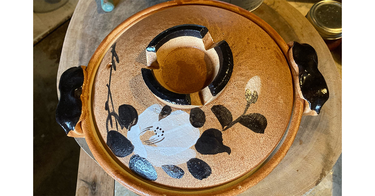 Photo of vintage donabe, a round lidded light brown ceramic bowl with dark brown handles and a matching lid painted with a white camelia flower with dark leaves.