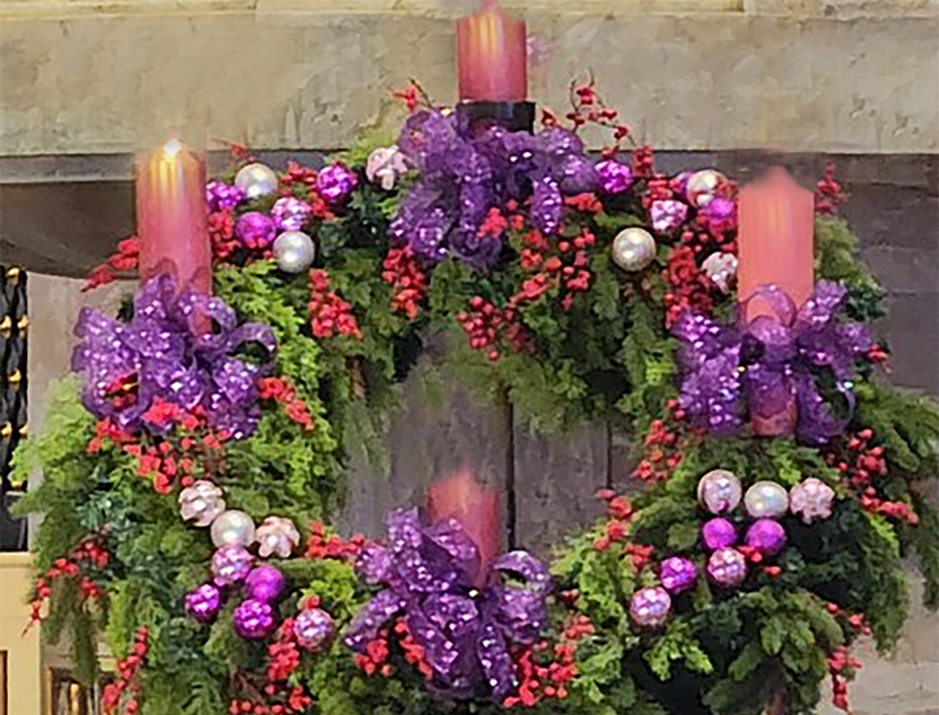 Wreath of conifer greenery and red and purple berries. Four candles, the left, top, and bottom candle are lavender in color, and the right hand one is pink. the left candle is lit.